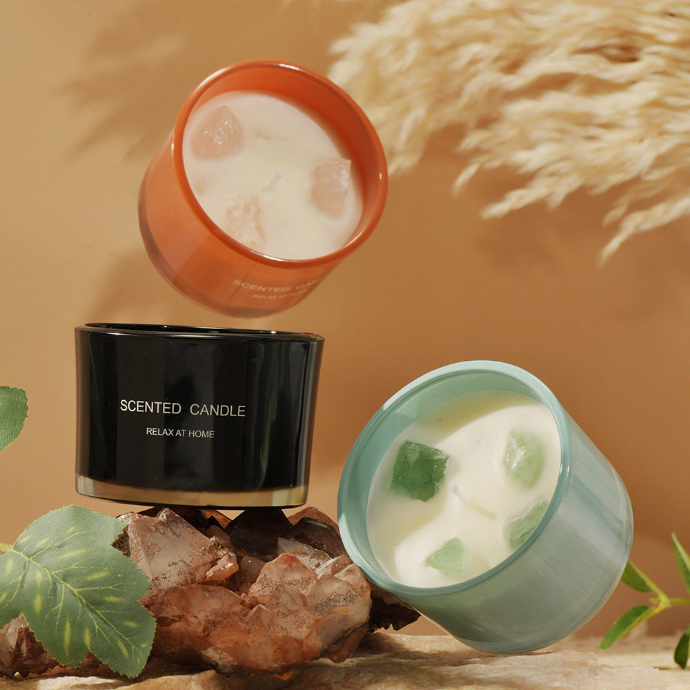 Three scented candles in different colors on a textured surface with plants and rocks.