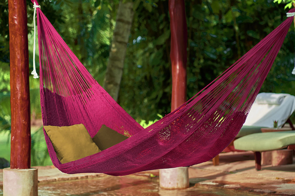 Pink hammock with green cushions hanging between two wooden poles in a garden setting.
