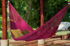 Pink hammock with green cushions hanging between two wooden poles in a garden setting.