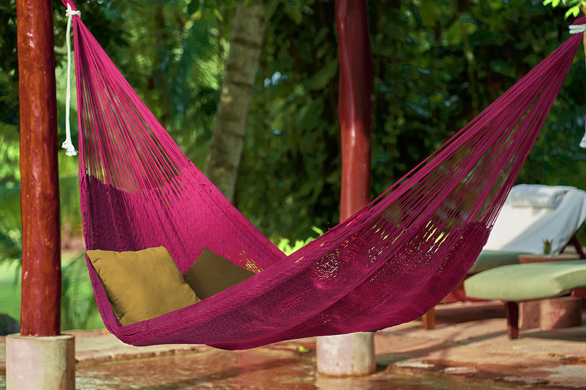 Pink hammock with green cushions hanging between two wooden poles in a garden setting.